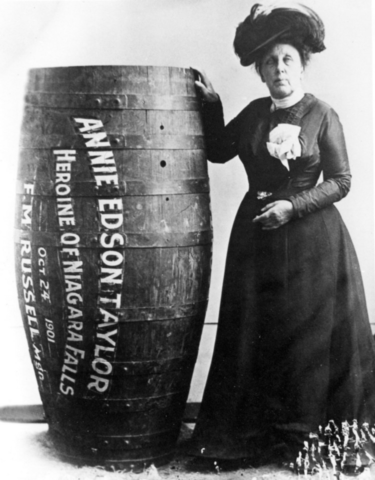 Annie Edson Taylor standing beside her oak barrel, which is labelled 'Annie Edson Taylor / Heroine of Niagara Falls / Oct 24 1901', in a formal black dress
