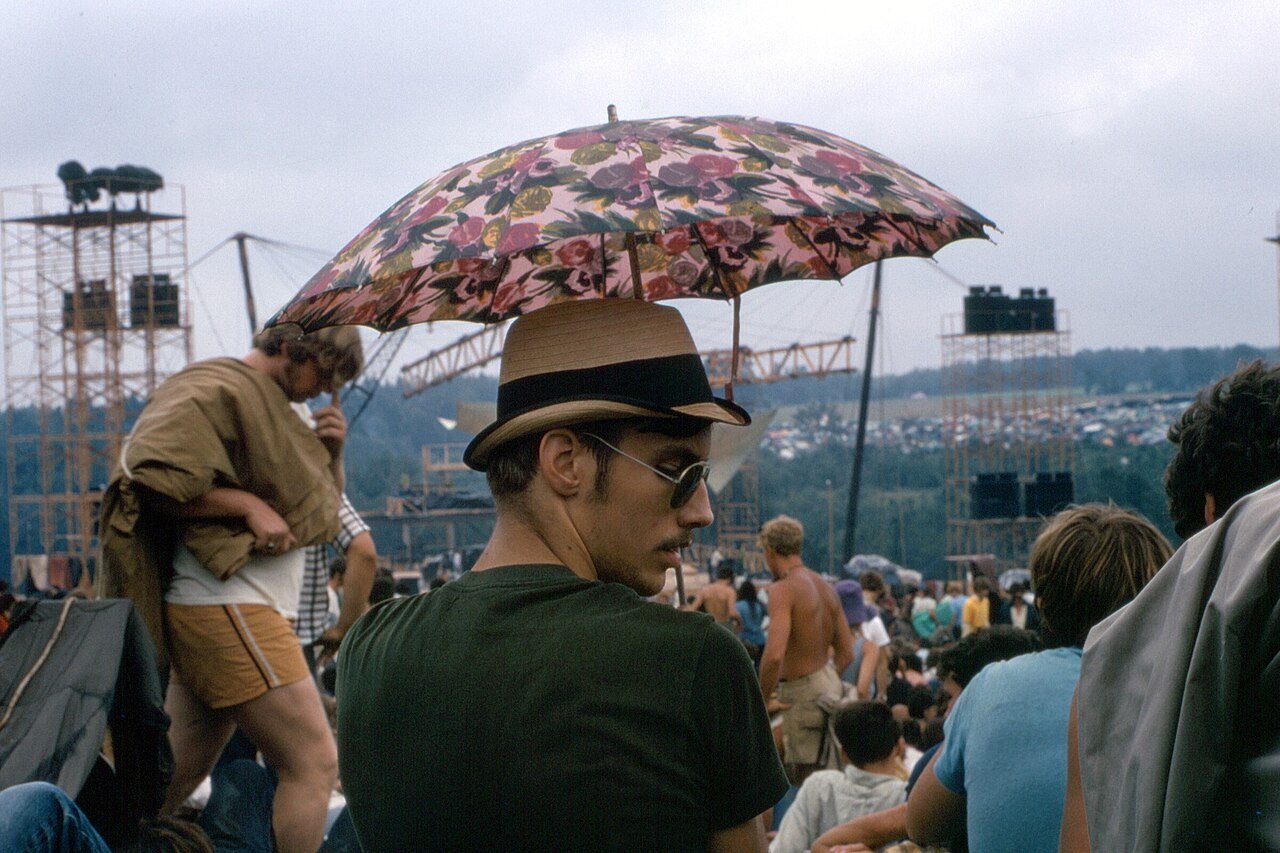 A festivalgoer under a parasol on the field, with the stage towers behind. Photo: James M Shelley, CC BY-SA 4.0, via Wikimedia Commons